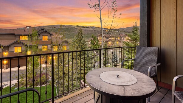 Varanda com mesa redonda e cadeiras, com vista para uma área residencial com casas e um pôr do sol.