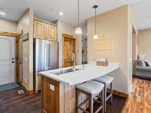 A kitchen with a central island, wooden cabinets, stainless steel appliances, pendant lights, and a gray sofa in the adjacent living area. - Mins to Park City & Deer Valley! 4BR Townhome (Hideout)