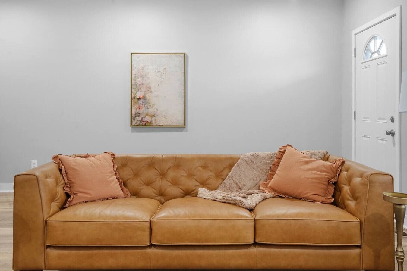 En beige lædersofa med flæset puder, et indrammet abstrakt maleri og en hvid dør med et vindue.