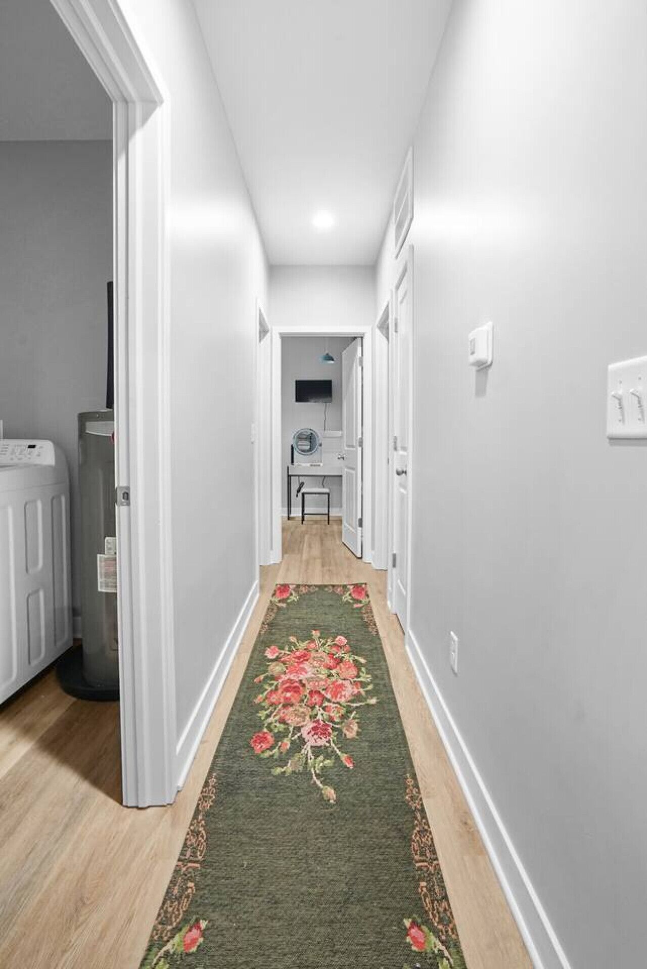 Um corredor com carpete floral, paredes brancas e uma porta que dá para um quarto com escrivaninha e cadeira.