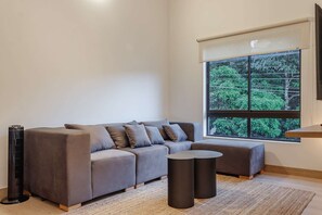A modern living room with a grey sectional sofa, a small round table, and a large window with a view of greenery. - NOK Peaceful 3BR in Medellin (Medellín)