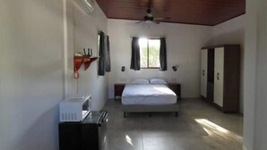 Ein Schlafzimmer mit Bett, Deckenventilator, Fenster mit Vorhängen, Kleiderschrank und einem kleinen Tisch mit einer Mikrowelle.
