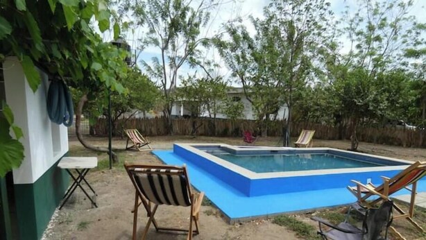 Ein rechteckiger Pool mit blauer Umrandung, umgeben von einer Sandfläche mit zwei Holzstühlen und einem Metallstuhl.