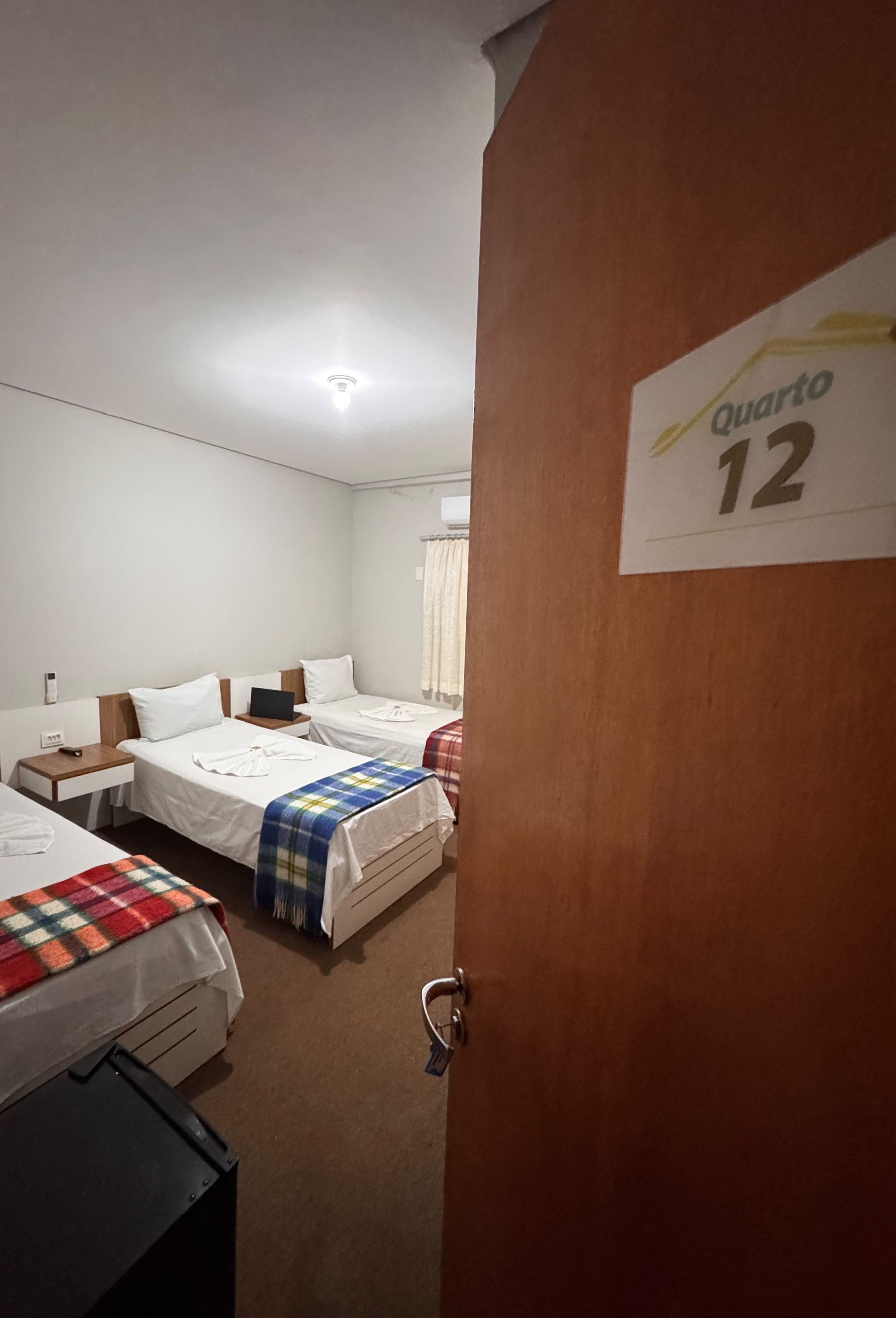Habitación de hotel con dos camas, un escritorio y una puerta con la leyenda 'Quarto 12'.