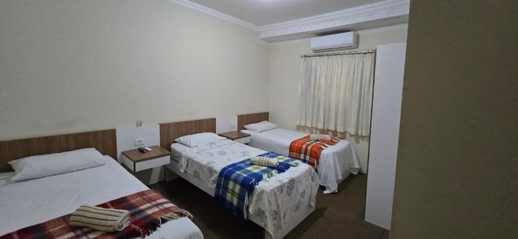 Una habitación de hotel con tres camas, una mesita de noche con lámpara, una ventana con cortinas y un equipo de aire acondicionado.