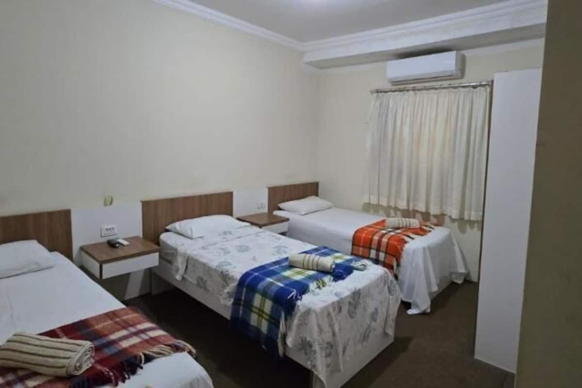 Una habitación de hotel con tres camas, una mesita de noche con lámpara, una ventana con cortinas y un equipo de aire acondicionado.