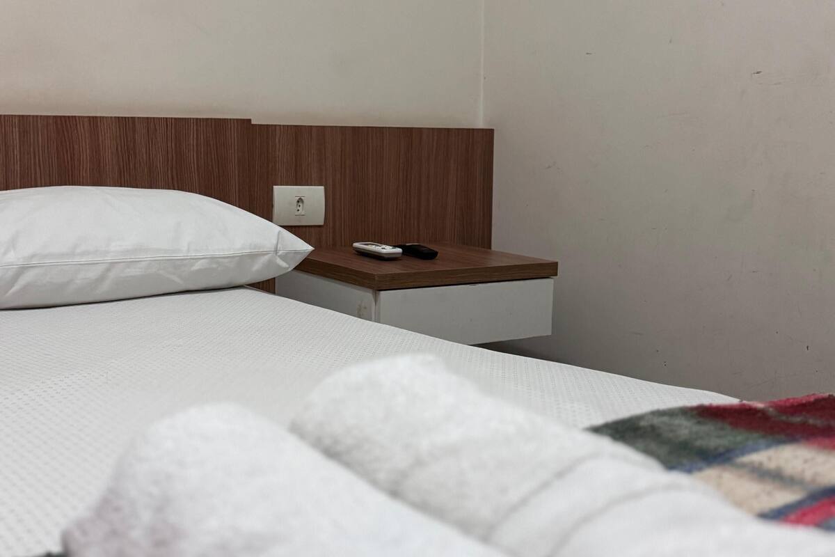Habitación de hotel con cama, cabecera de madera, mesita de noche con teléfono y control remoto, y toallas enrolladas sobre la cama.