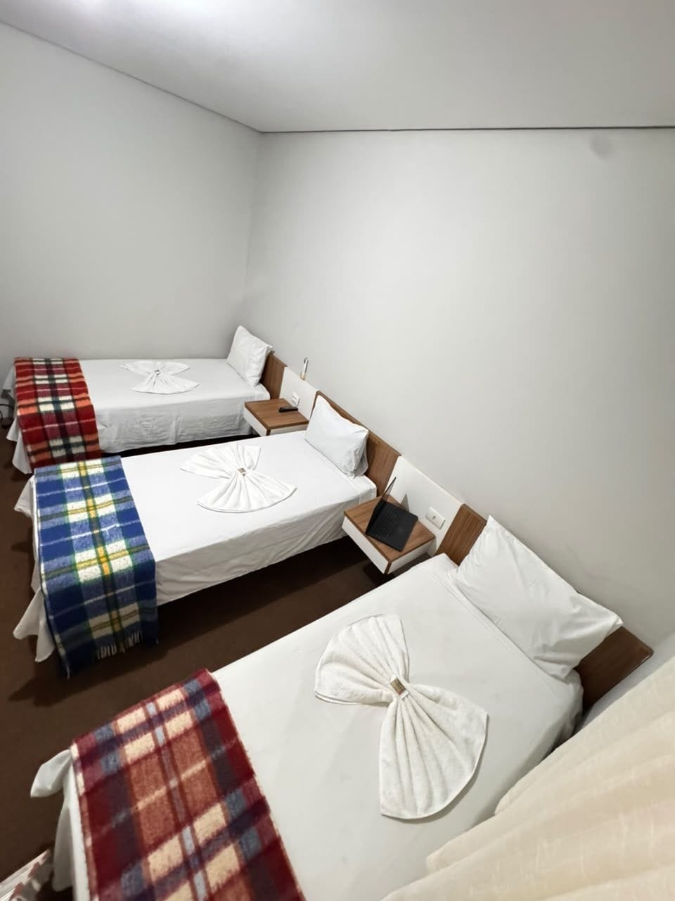 Una habitació d'hotel amb quatre llits individuals, cadascun amb un llençol blanc i un coixí decoratiu. Hi ha una taula de nit de fusta amb un portàtil sobre un dels llits.