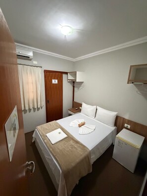 A hotel room with a bed, bedside tables, a desk, a lamp, an air conditioning unit, and a door with a sign. - HOTEL NORTE SUL (Campo Grande)