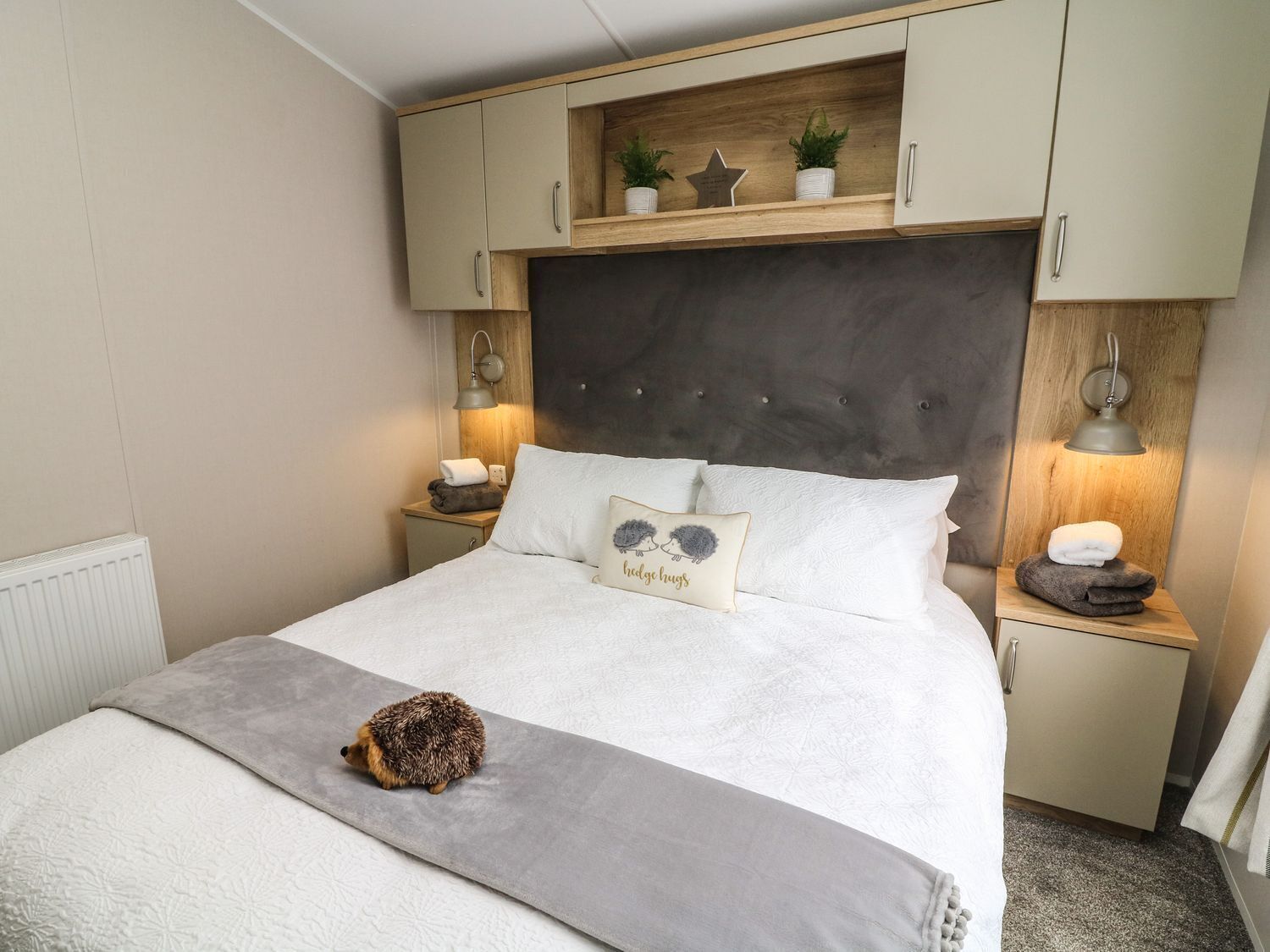 Un dormitorio con cama, mesita de noche, lámpara, estante con adornos y un pequeño erizo sobre la cama.