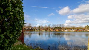 A calm lake with a row of cabins and a dense green bush in the foreground. - Hedgehog Lodge (Carnforth)