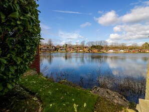 A calm lake with a row of cabins and a dense green bush in the foreground. - Hedgehog Lodge (Carnforth)