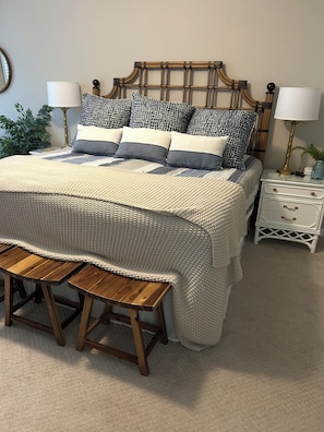 Un lit bien fait, recouvert d’une couvre-lit à motifs, un banc en bois au pied du lit et une table de chevet avec une lampe et une petite plante.