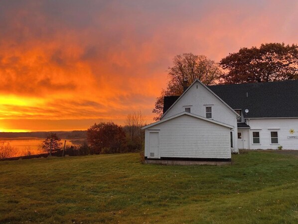 A white building with a garage, a small window, and a door, located next to a body of water with a sunset in the background.