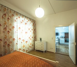 Ein Schlafzimmer mit Bett, Schminktisch, einer Zimmerpflanze und Blick zur Küche.