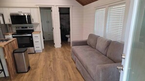 Un espacio habitable compacto con kitchenette, un sofá gris y una puerta que da a un baño.
