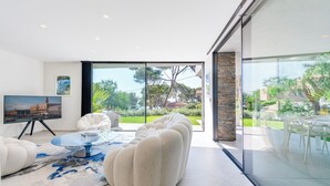 Een moderne woonkamer met een grote glazen deur die uitkomt op een tuin, een ronde salontafel en een comfortabele bank.