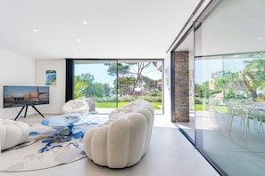Ein modernes Wohnzimmer mit einem großen Glasfenster, das in einen Garten führt, einem runden Couchtisch und einer gemütlichen Couch.