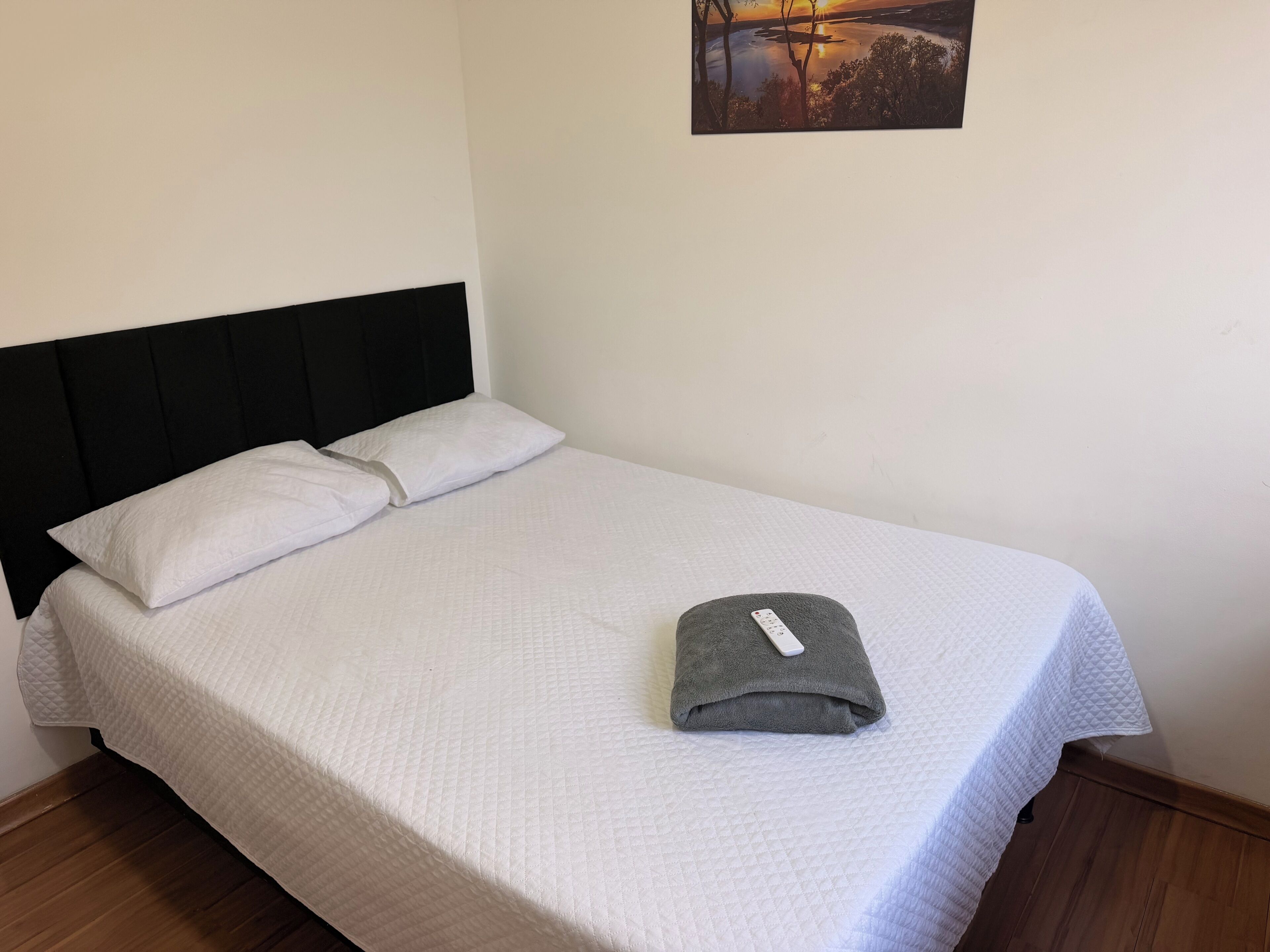 Un letto rifatto con una trapunta bianca, un asciugamano grigio piegato e una testiera nera.