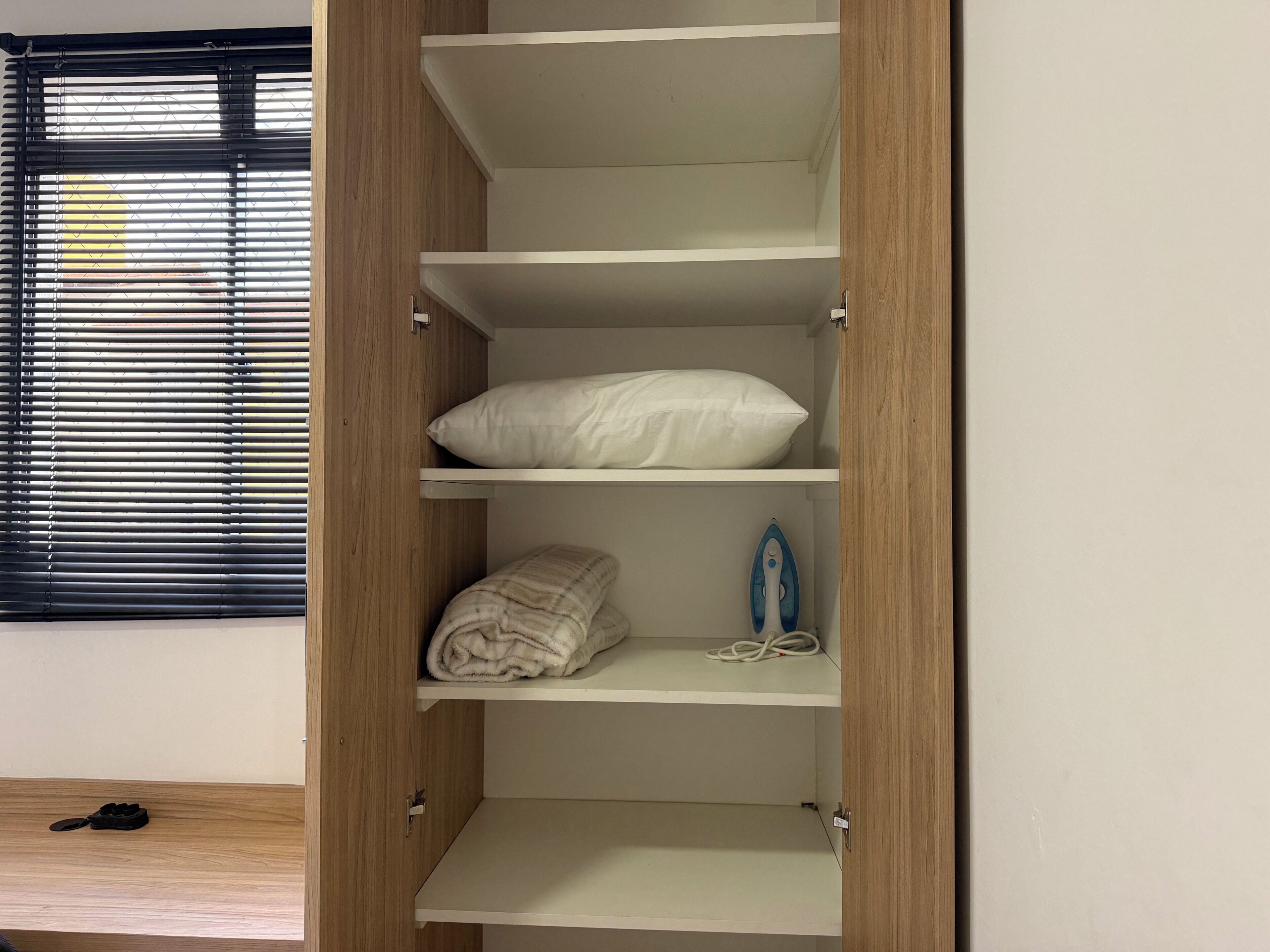 A room with a wooden wardrobe, a window with blinds, and a pair of shoes on the floor.