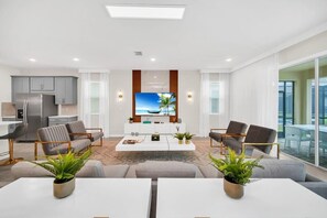 Una sala de estar moderna con un televisor de pantalla plana, sofás grises, una mesa de centro y un comedor con una mesa y sillas blancas.