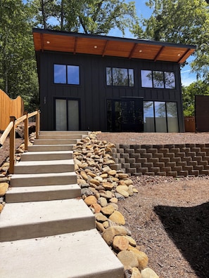 Un bâtiment moderne avec un escalier en béton menant à une porte en bois, le tout entouré d’un mur de pierre et d’une clôture en bois.