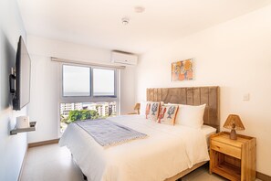 A bedroom with a bed, a nightstand, a wall-mounted TV, a window, and an air conditioning unit. - Ocean View Condo Steps to Beach & Comfort (Jacó)