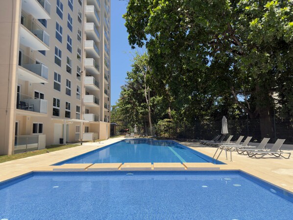 A swimming pool with a tiled deck, surrounded by trees and a multi-story building with balconies. - Ocean View Condo Steps to Beach & Comfort (Jacó)