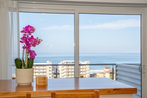 A balcony with a wooden table, a potted orchid, and a view of the ocean and buildings. - Ocean View Condo Steps to Beach & Comfort (Jacó)