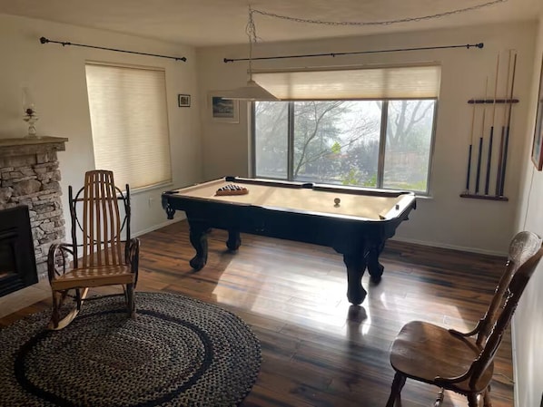 A pool table in a room with a fireplace, a large window, and a view of trees.