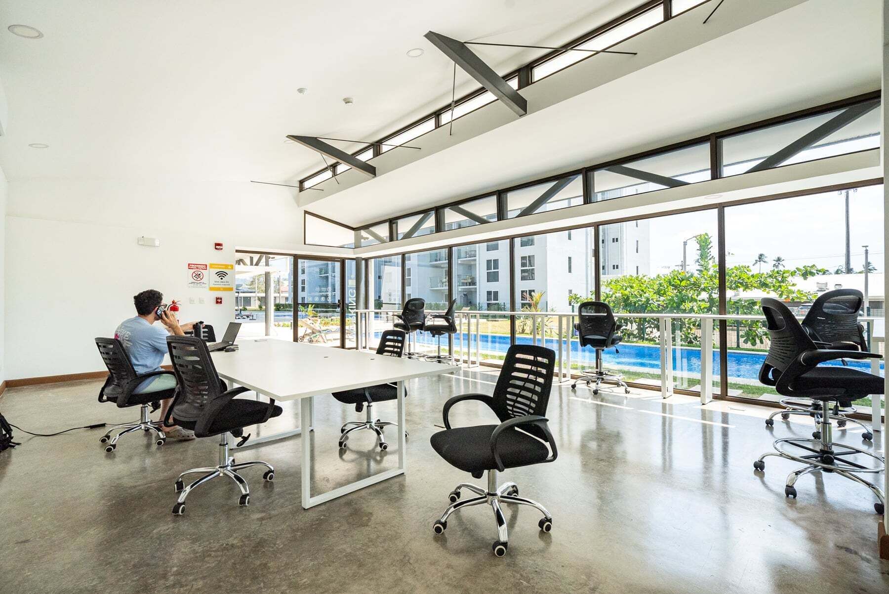 A modern conference room with a glass table, black office chairs, and large windows offering a view of a swimming pool and buildings.