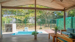 Un espacio exterior cubierto con piscina, muebles de madera y vista a un jardín cercado.