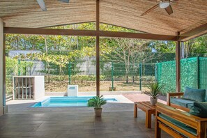 A covered outdoor area with a pool, wooden furniture, and a view of a fenced garden. - Private Pool & Tropical Style, Minutes From the Beach (Jacó)