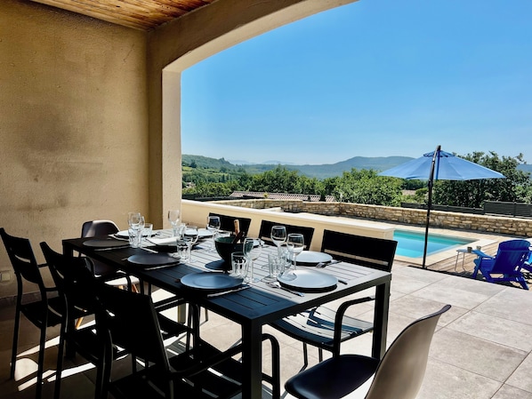 Un espace de restauration en plein air avec une table dressée pour quatre personnes, offrant une vue sur une piscine et un paysage pittoresque.