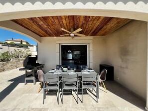 Eine Terrasse im Freien mit Tisch für vier Personen, Deckenventilator und Grill.