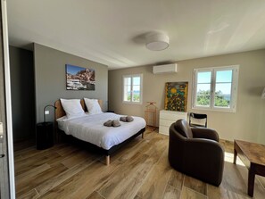 Ein Schlafzimmer mit einem Bett, einem Sessel, einem Tisch, einer Kommode und zwei Fenstern.