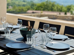 Une table dressée pour le repas, avec de la vaisselle et des verres transparents, une cuillère en bois dans un bol, et en arrière-plan, une vue sur la verdure et des bâtiments.
