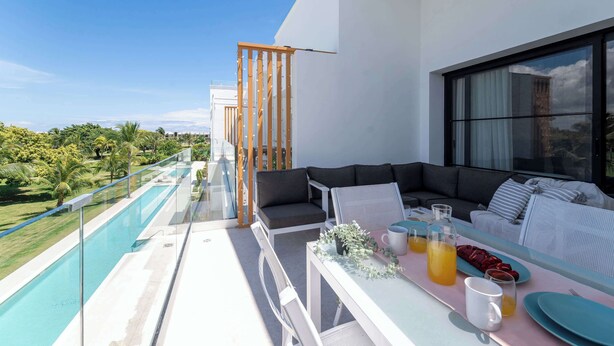 Ein Balkon mit Glasgeländer, einer Couch, einem Tisch mit Getränken und Tellern sowie Blick auf Grünflächen und einen Pool.