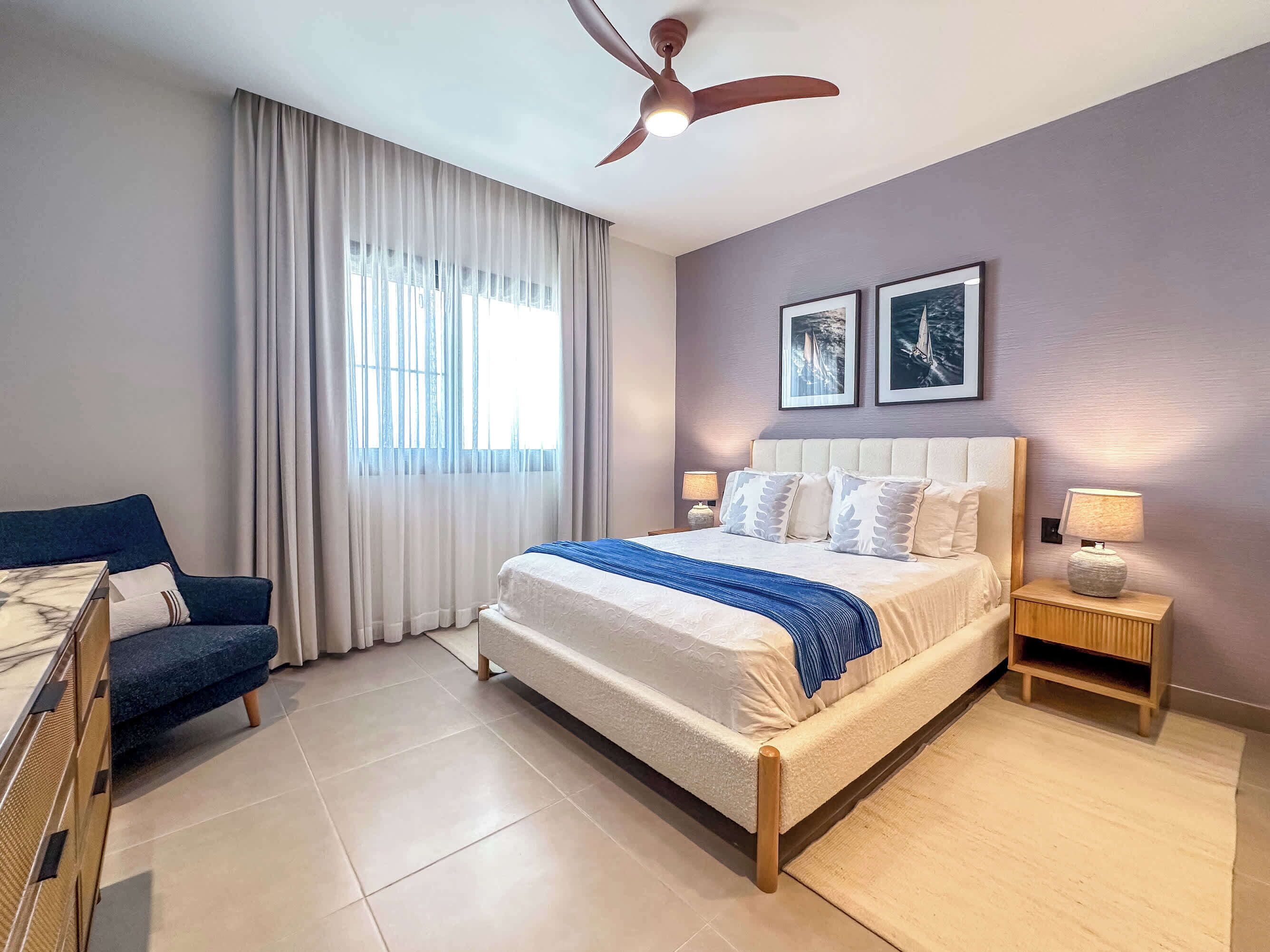 Habitación de hotel con cama, escritorio, silla, ventilador de techo y dos cuadros en la pared.