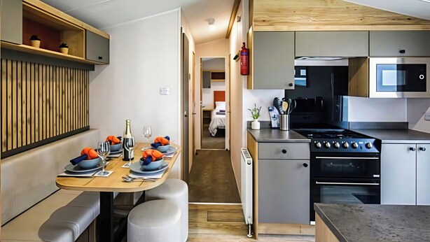 A compact kitchen with modern appliances, a dining area with a bench, and a visible hallway leading to a bedroom. - 26 Ocean View (Llwyngwril)