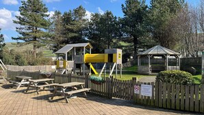 A playground with a wooden fence, picnic tables, and a gazebo. - 26 Ocean View (Llwyngwril)