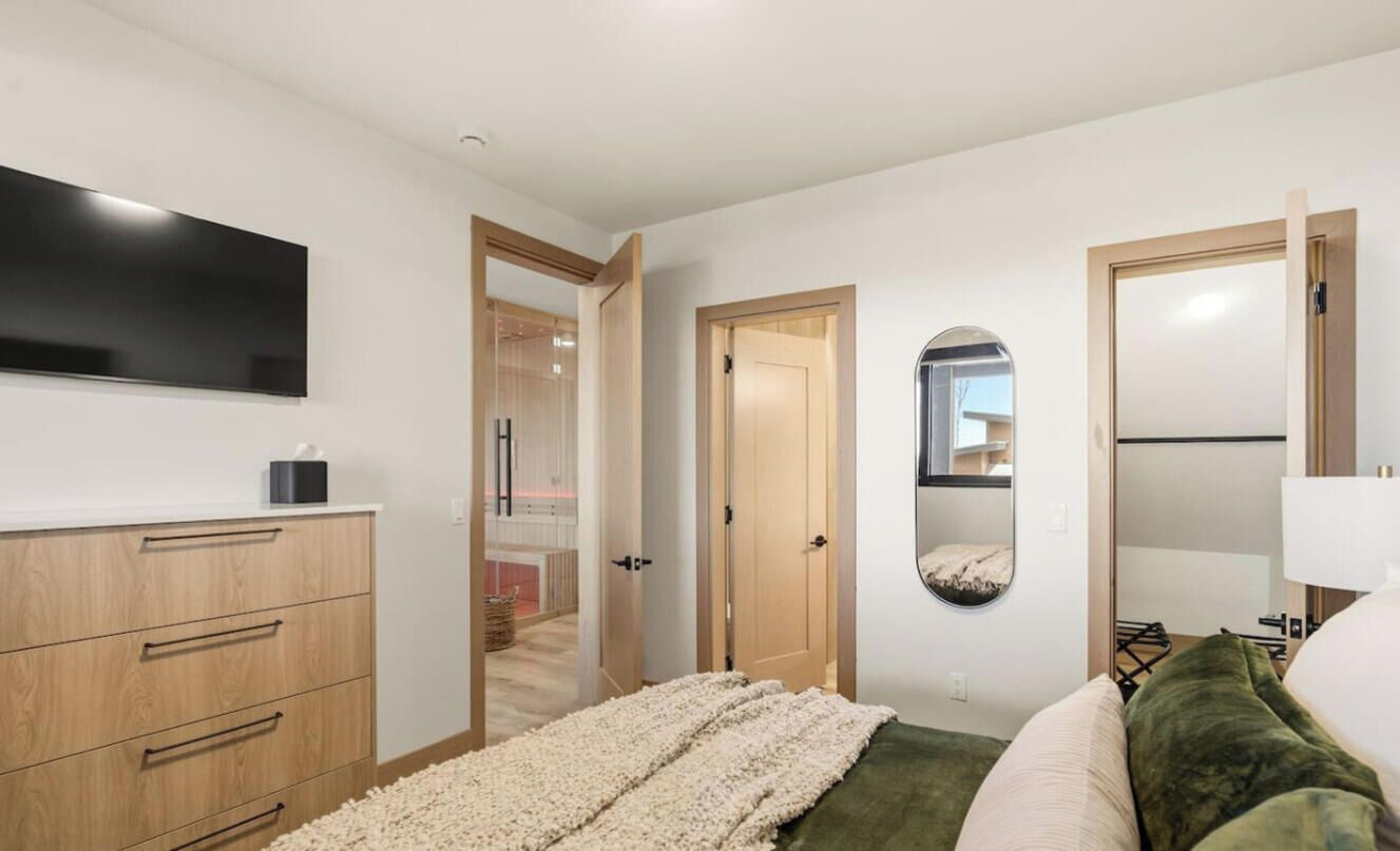 A bedroom with a bed, a TV, a dresser, a mirror, and a door leading to a bathroom.