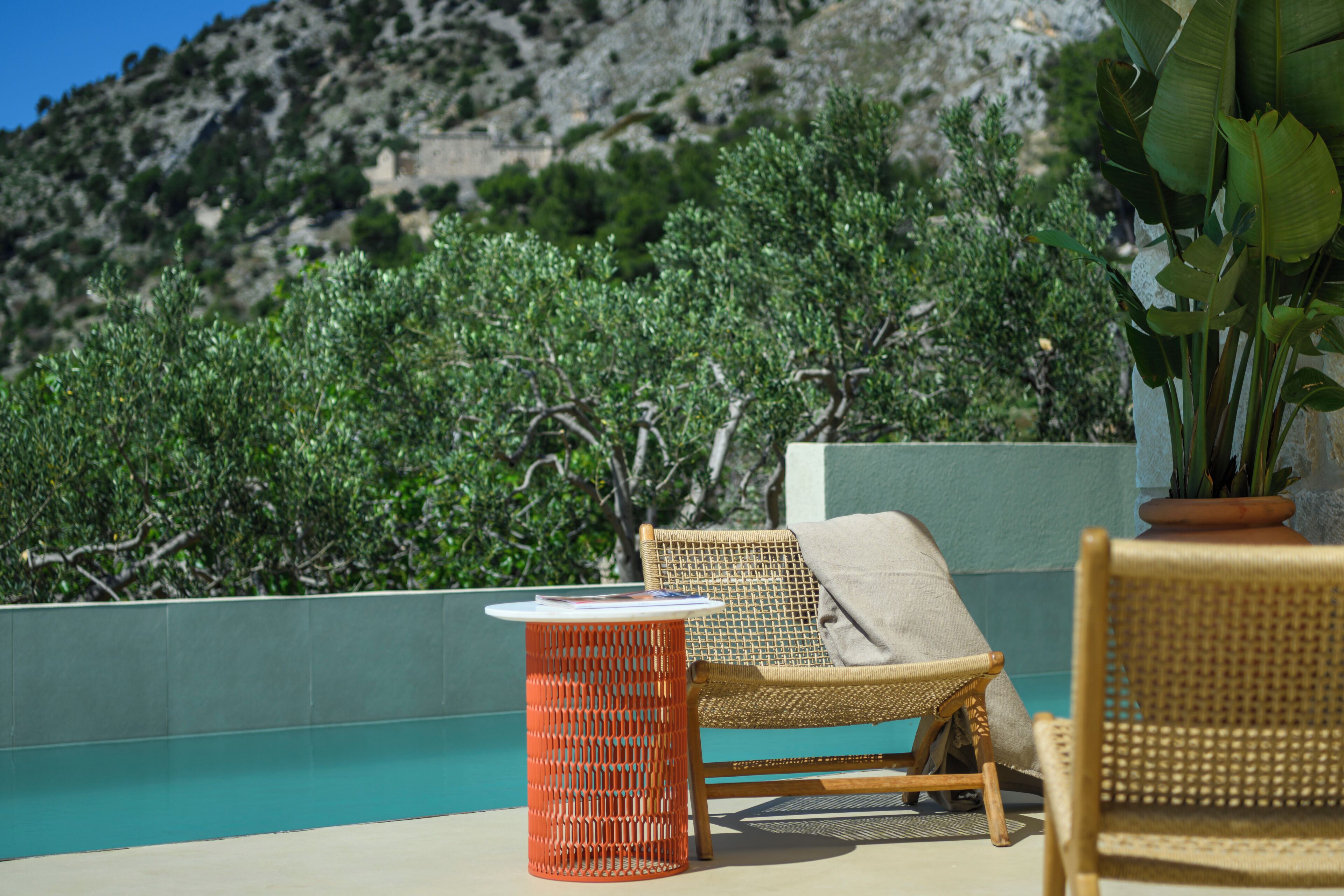 A wicker lounge chair with a cushion, a small orange table, and a potted plant on a terrace with a pool and a view of a mountainous landscape.