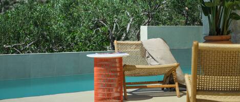 A wicker lounge chair with a cushion, a small orange table, and a potted plant on a terrace with a pool and a view of a mountainous landscape.