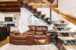 Una sala de estar moderna con un sofá de cuero, una escalera con escalones de madera y barandilla de metal, y una cocina al fondo.