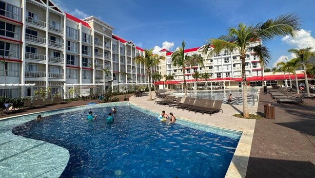 Zona de piscina de hotel con una alberca grande, camastros y un edificio con balcones.