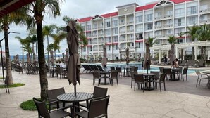 Zona de piscina hotelera con mobiliario exterior y un edificio de varios pisos al fondo.