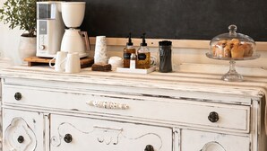 Une commode blanche avec des tiroirs, une machine à café, un bocal en verre et un tableau noir posés sur une surface en bois.
