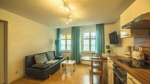 Ein kompakter Wohnraum mit Sofa, Esstisch, Küchenzeile mit Geräten und einem an der Wand befestigten Fernseher.