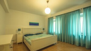 Ein Schlafzimmer mit einem Bett, Nachttischen, einer Stehlampe, einer Deckenleuchte, einem Fenster mit Vorhängen und einem Wandbild.